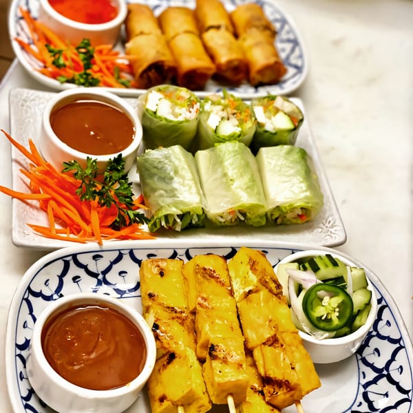 Plates of Thai spring rolls, fresh rolls, and chicken satay with dipping sauces and garnishes.