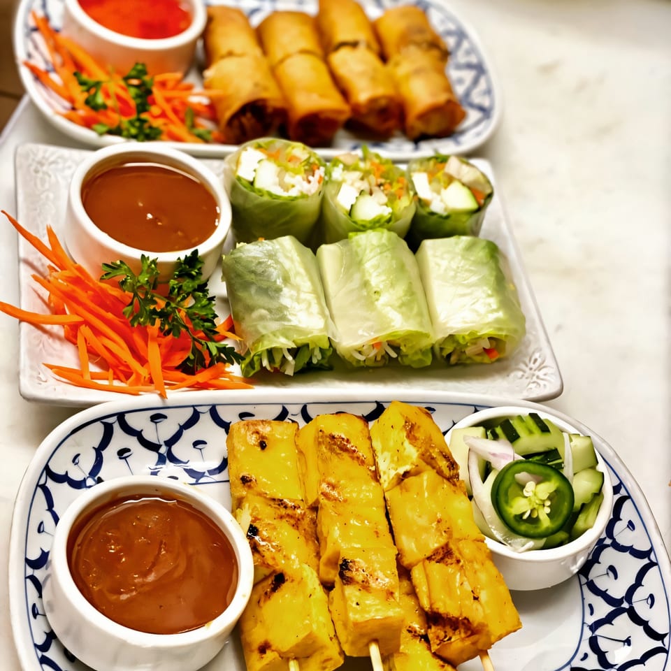 Plates of Thai spring rolls, fresh rolls, and chicken satay with dipping sauces and garnishes.