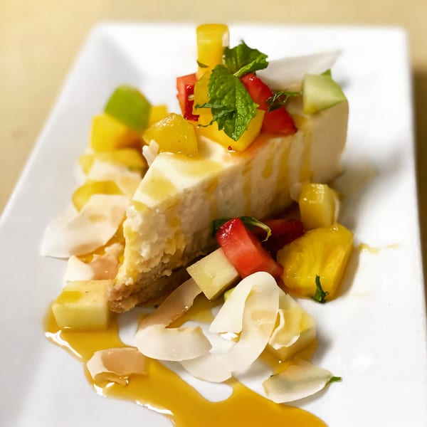 Slice of cheesecake topped with mango, strawberries, mint, and coconut flakes, drizzled with sauce.