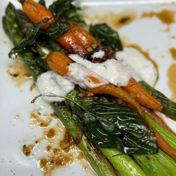 Roasted carrots and asparagus with a creamy white sauce and basil leaves on a white plate.